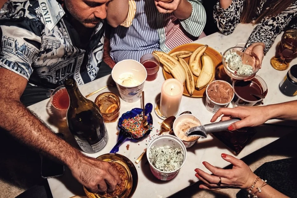 Overhead shot of a group enjoying ice cream and bananas at a white marble table with colorful sprinkles, drinks, and candlelight.