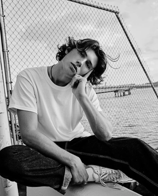 Black and white portrait of a young adult man with dark hair, wearing a white shirt, seated in front of a wire fence and body of water.