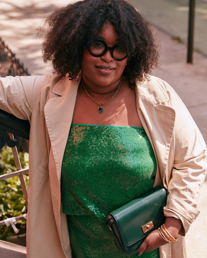 A stylish Black woman poses outdoors in a green sequin dress and trench coat, holding a clutch, showcasing fashion and street style.