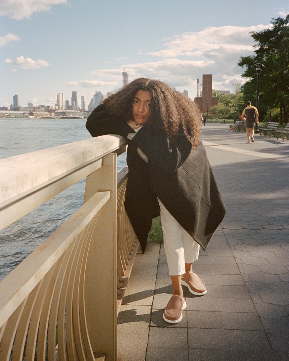 Woman with curly hair leaning against a railing on a waterfront walkway with a cityscape backdrop on a sunny day, medium shot.