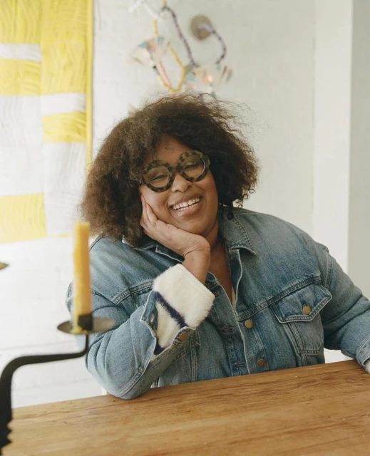 Portrait of a smiling woman with glasses wearing a denim jacket, seated at a table in a brightly lit interior, with colorful artwork.