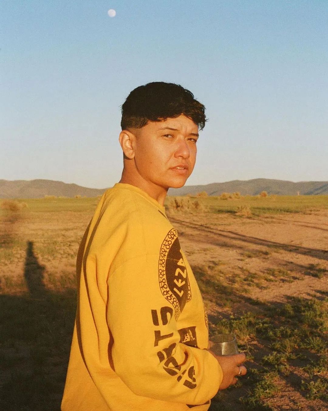 Portrait of an adult with short brown hair wearing a yellow sweatshirt outside in a field with hills and blue sky with a moon.