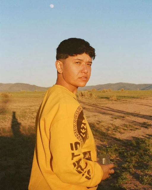 Portrait of an adult with short brown hair wearing a yellow sweatshirt outside in a field with hills and blue sky with a moon.