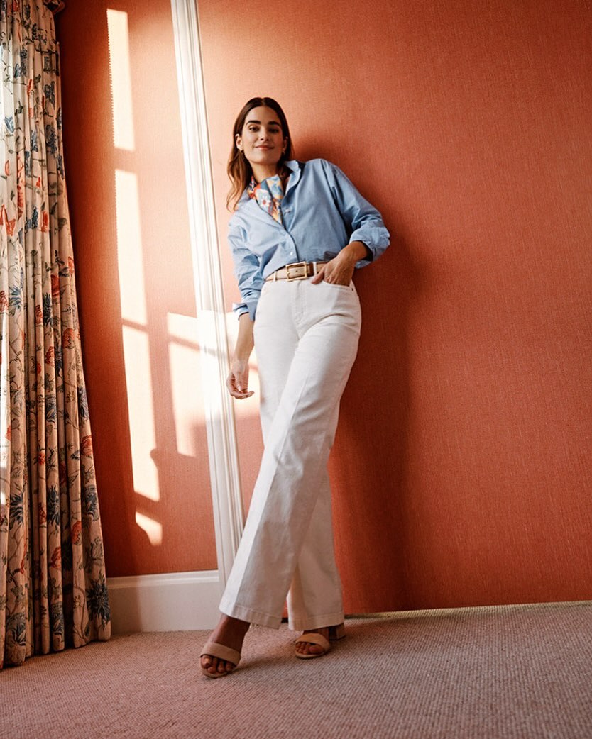 Adult woman in casual fashion stands indoors against a salmon-colored wall, wearing a light blue button-up shirt and white wide-leg pants.