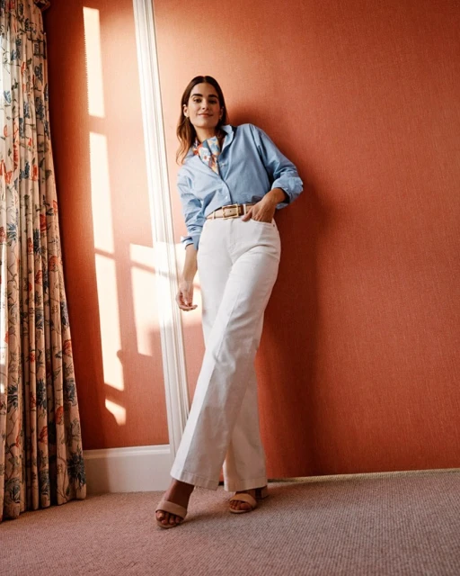Adult woman in casual fashion stands indoors against a salmon-colored wall, wearing a light blue button-up shirt and white wide-leg pants.