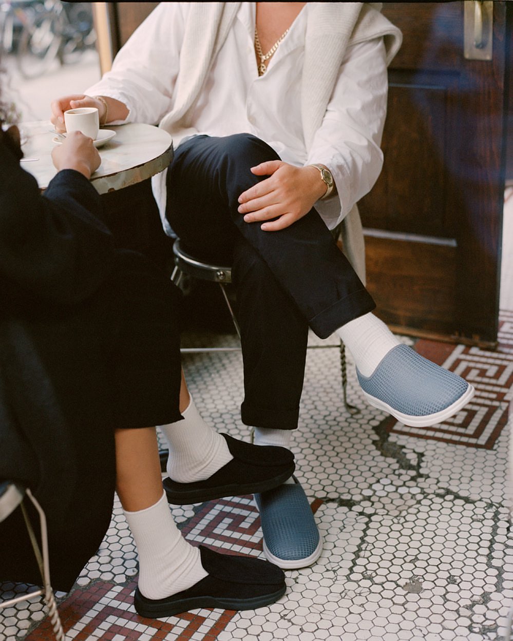 Two people sitting at a cafe table, showcasing black and light blue slip-on shoes with white socks on a patterned tile floor.