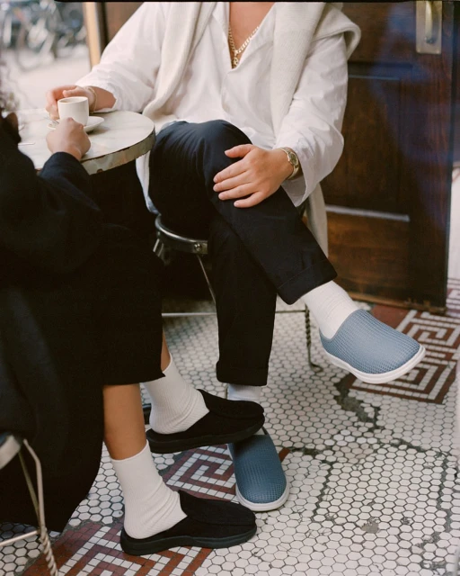 Two people sitting at a cafe table, showcasing black and light blue slip-on shoes with white socks on a patterned tile floor.