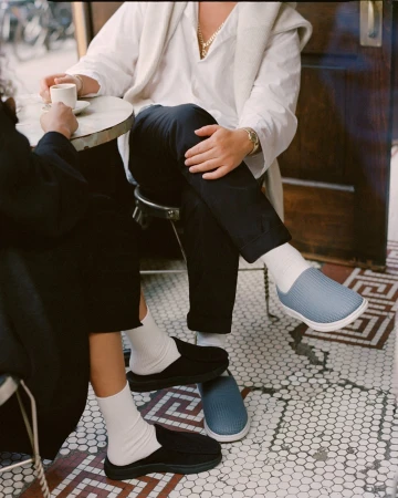 Two people sitting at a cafe table, showcasing black and light blue slip-on shoes with white socks on a patterned tile floor.