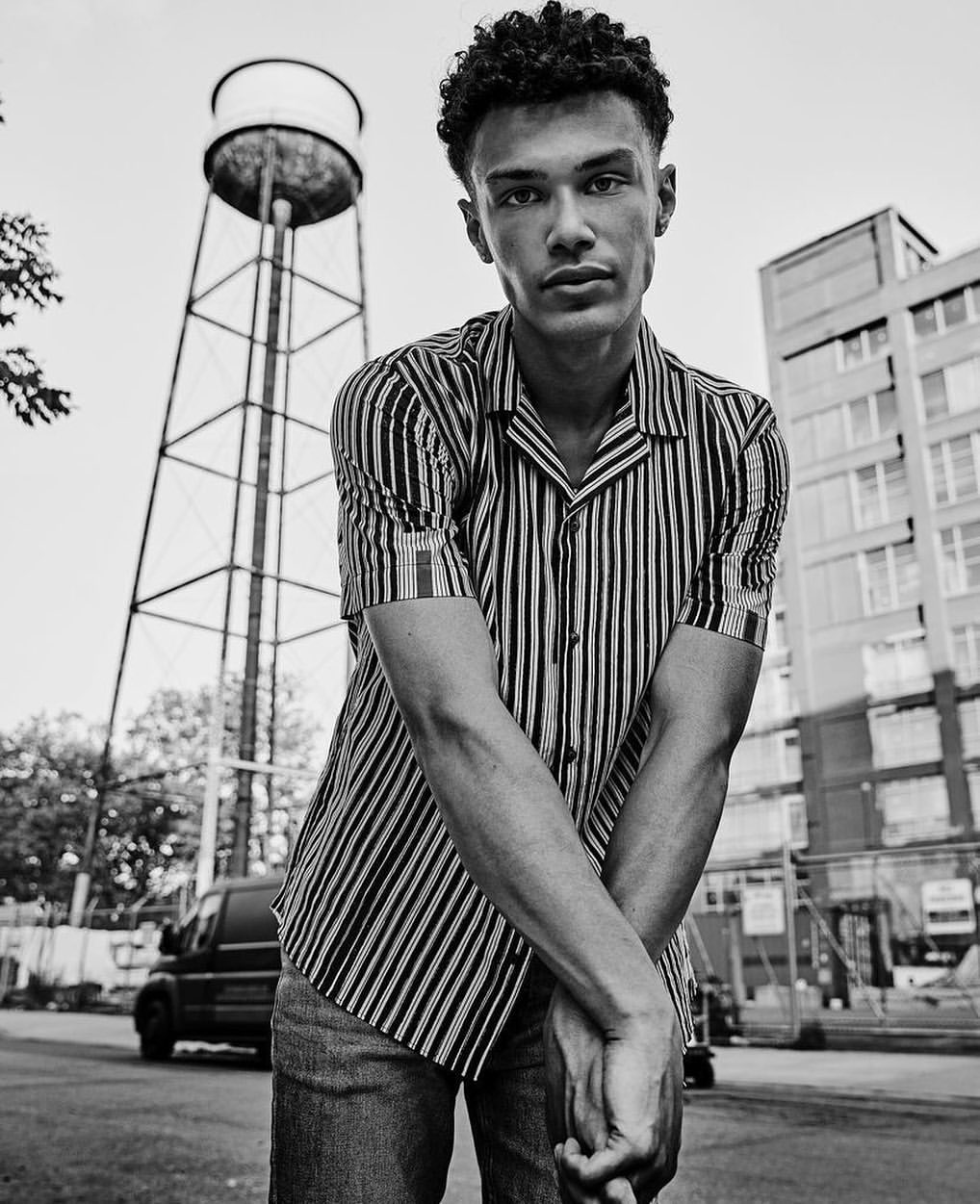 Portrait of a young adult male in striped shirt and jeans, posing outdoors with a water tower and urban buildings in the background.