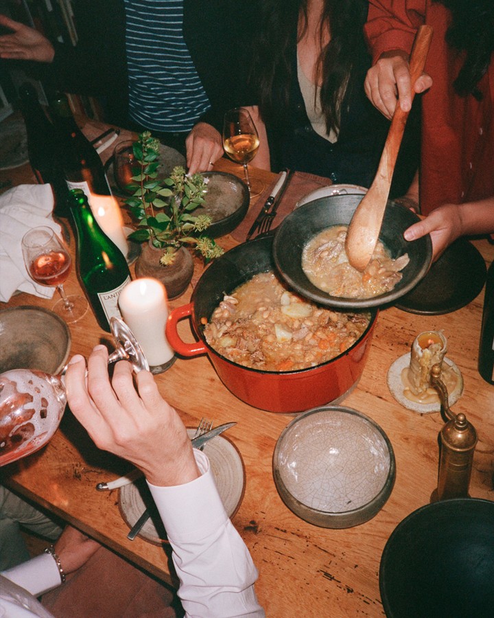 Overhead shot of a communal table with people serving stew, wine glasses, plates, candles, and a plant, evoking a cozy gathering.