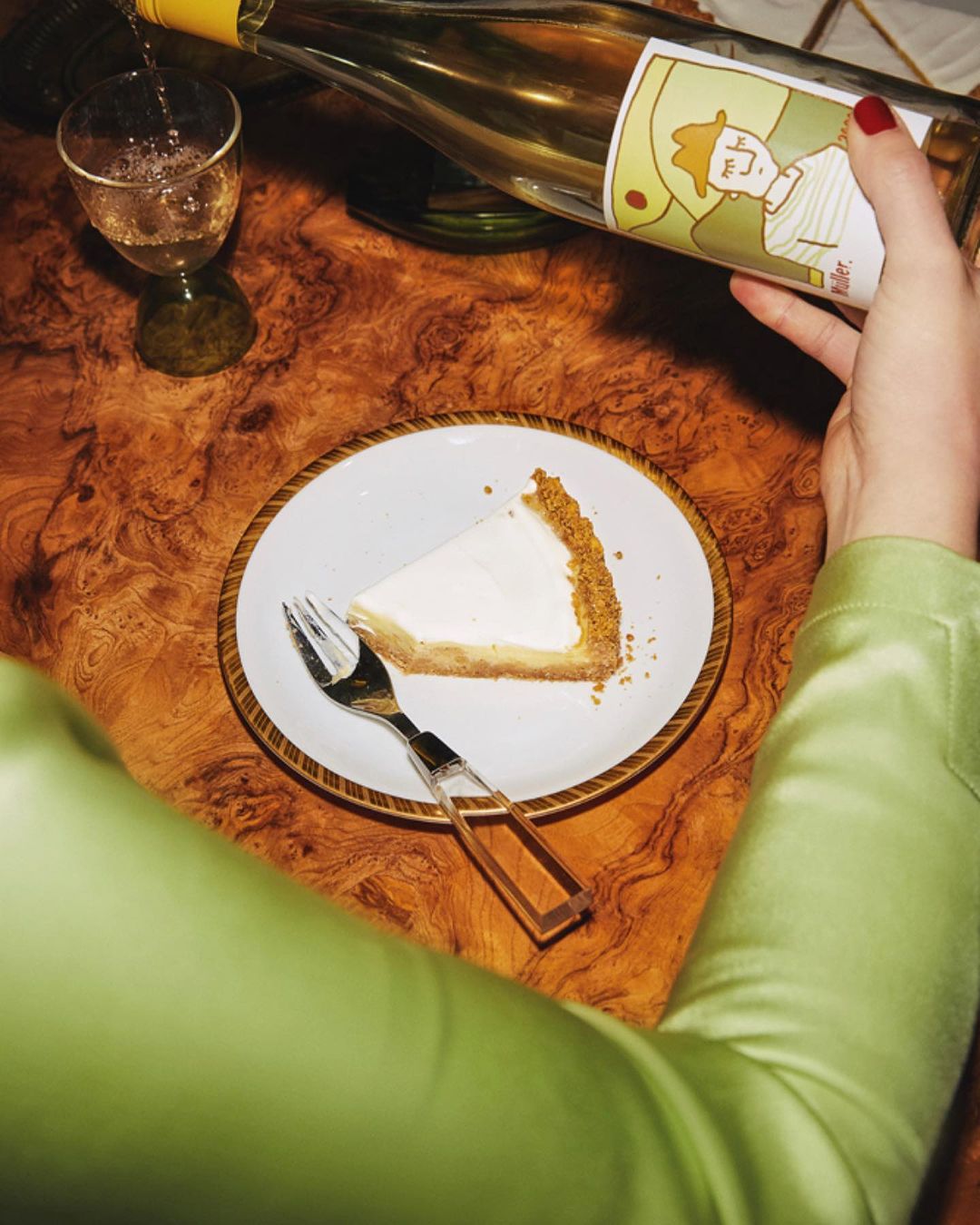 Overhead shot of a slice of cheesecake on a white plate, a fork, and a bottle of white wine being poured into a glass.