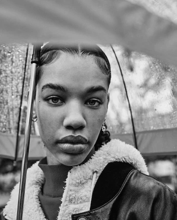 Black and white portrait of a young woman with an umbrella, wearing a jacket with a shearling collar in an outdoor setting.