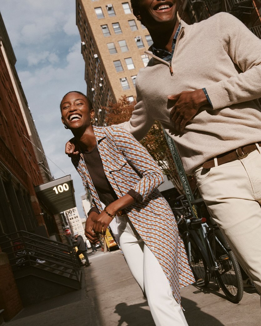 Two smiling people walk on a city sidewalk in daytime, with tall buildings, brownstones, and a bicycle in the background on a sunny day.