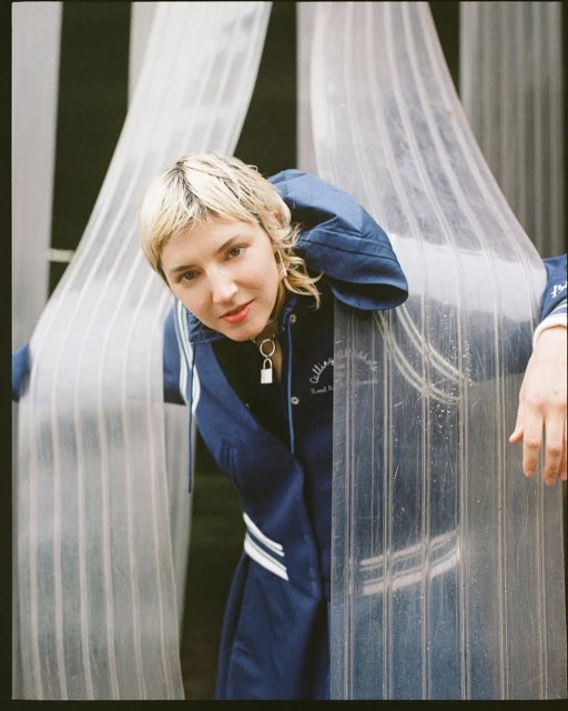 Stylish woman in a navy track jacket posing in front of a transparent, corrugated plastic sheet backdrop in a medium, eye-level shot.