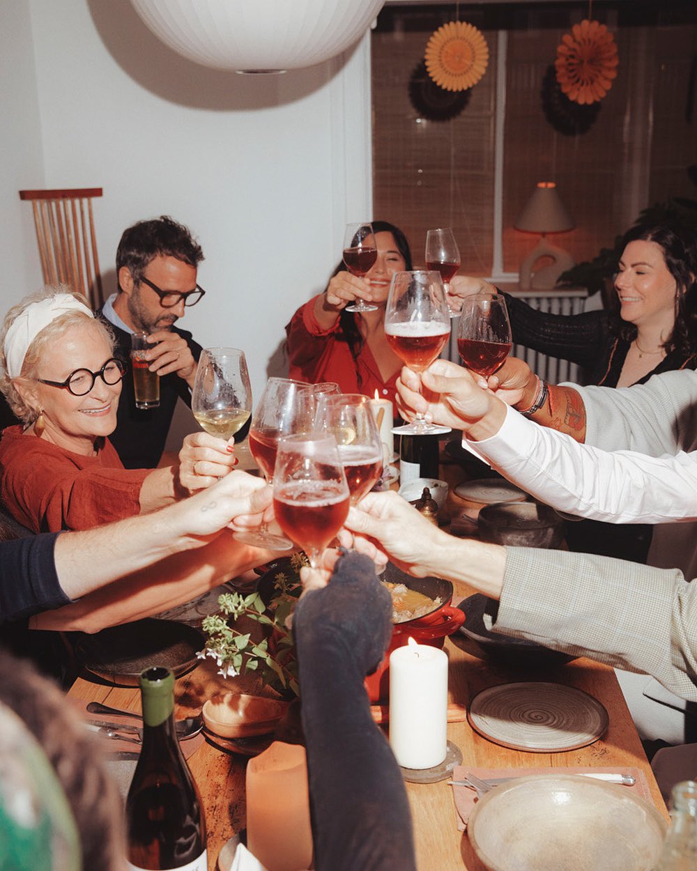A group of diverse adults gather indoors at a wood table toasting with wine glasses, creating a convivial social scene at home.