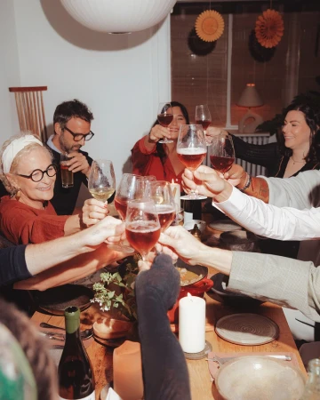 A group of diverse adults gather indoors at a wood table toasting with wine glasses, creating a convivial social scene at home.