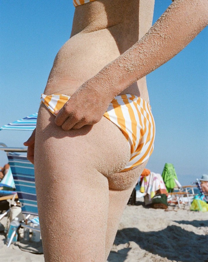 Woman on a beach with tan skin and yellow and white striped bikini bottom, covered in sand in a side view with a beach background.