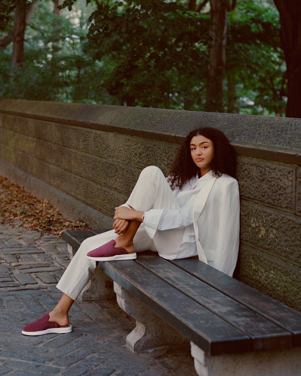 Woman in a white suit and burgundy mules sitting on a bench in an outdoor park setting with green foliage in the background.