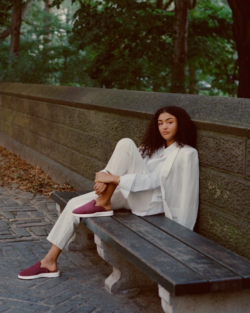 Woman in a white suit and burgundy mules sitting on a bench in an outdoor park setting with green foliage in the background.