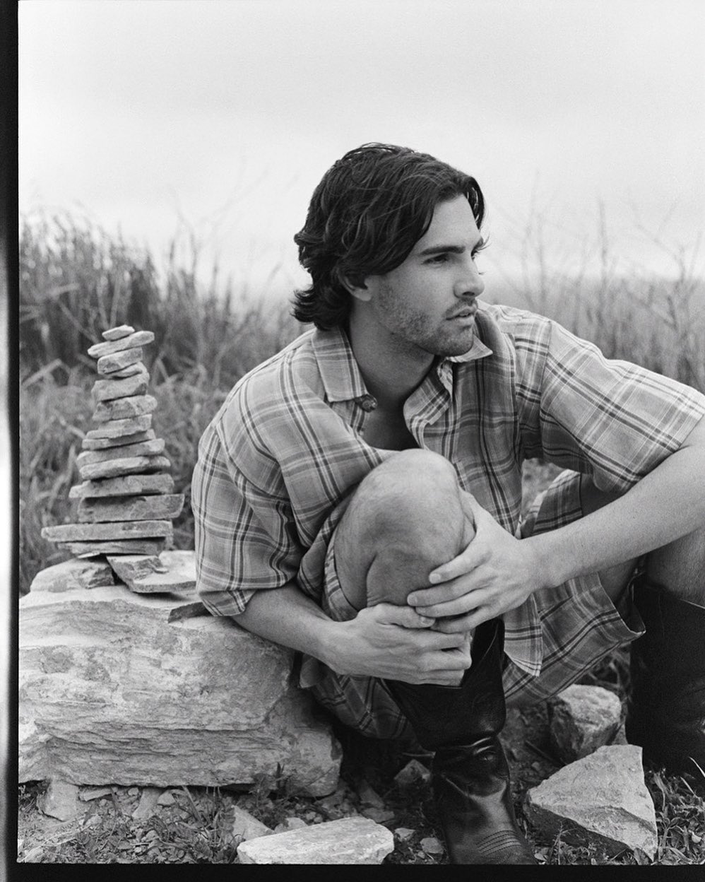 Black and white outdoor portrait of a man in a plaid shirt, sitting near a stone tower, with a grassy background under a cloudy sky.