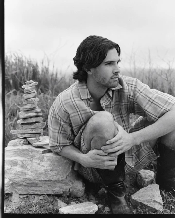 Black and white outdoor portrait of a man in a plaid shirt, sitting near a stone tower, with a grassy background under a cloudy sky.