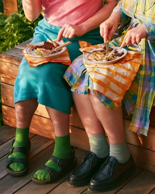Two people seated, eating outdoors on a wooden surface, with plates of food and napkins, showcasing a casual dining scene.