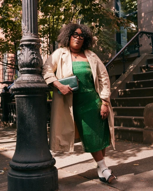 Adult woman in trench coat and green dress poses on city street next to black lamp post on sunny day, holding purse, looking at camera.