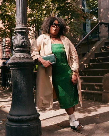 Adult woman in trench coat and green dress poses on city street next to black lamp post on sunny day, holding purse, looking at camera.