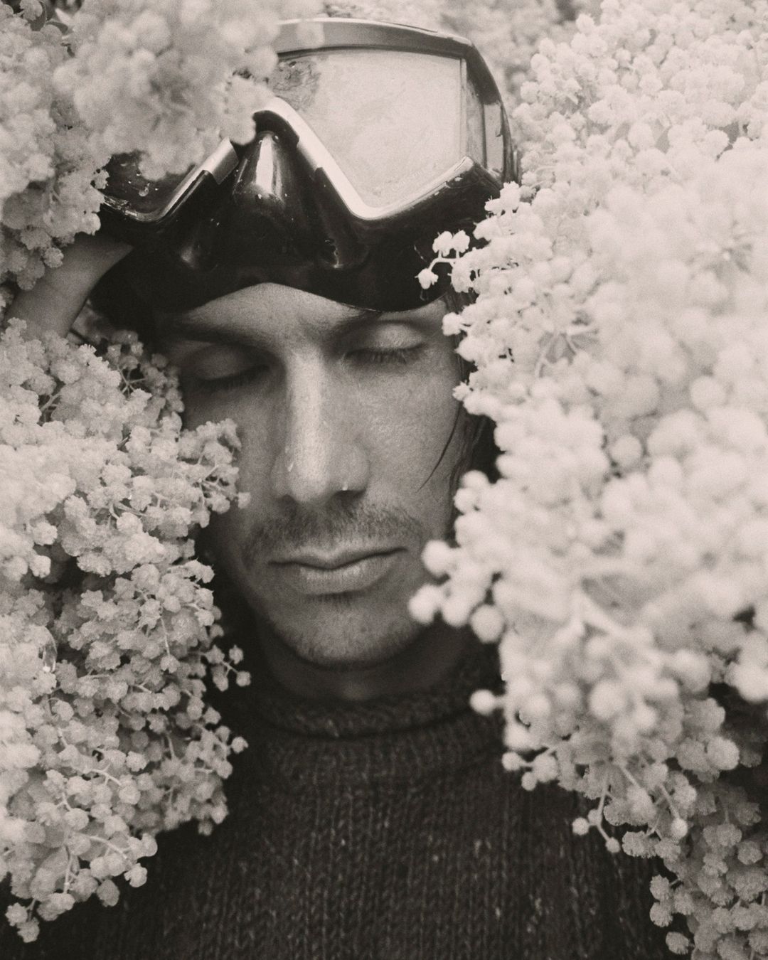 Monochromatic close-up shot of a man with closed eyes wearing ski goggles surrounded by clusters of small, white flowers.