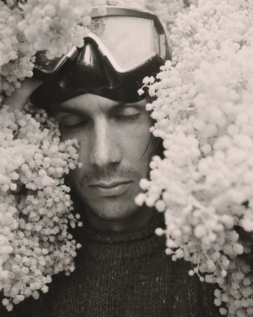 Monochromatic close-up shot of a man with closed eyes wearing ski goggles surrounded by clusters of small, white flowers.