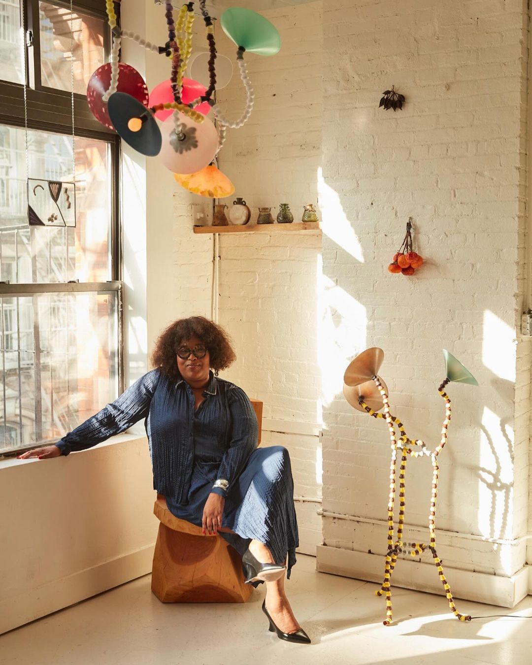 Woman with glasses wearing a blue outfit sitting on a brown chair in an airy room with artwork, and light fixtures. Interior decor.