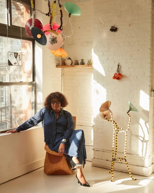 Woman with glasses wearing a blue outfit sitting on a brown chair in an airy room with artwork, and light fixtures. Interior decor.