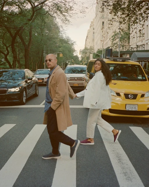 Couple walks hand-in-hand across a zebra crossing in an urban setting with yellow taxi cabs and tall buildings visible in the background.