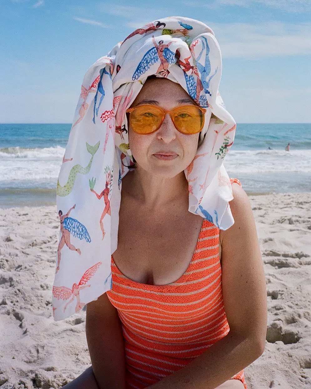 Woman in orange swimsuit and headscarf with mermaid print sits on a sunny beach with the ocean and blue sky visible in the background.