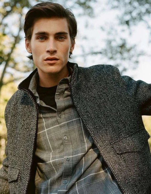 Man in herringbone coat and plaid shirt poses outdoors, showcasing classic menswear fashion against a blurred natural background.
