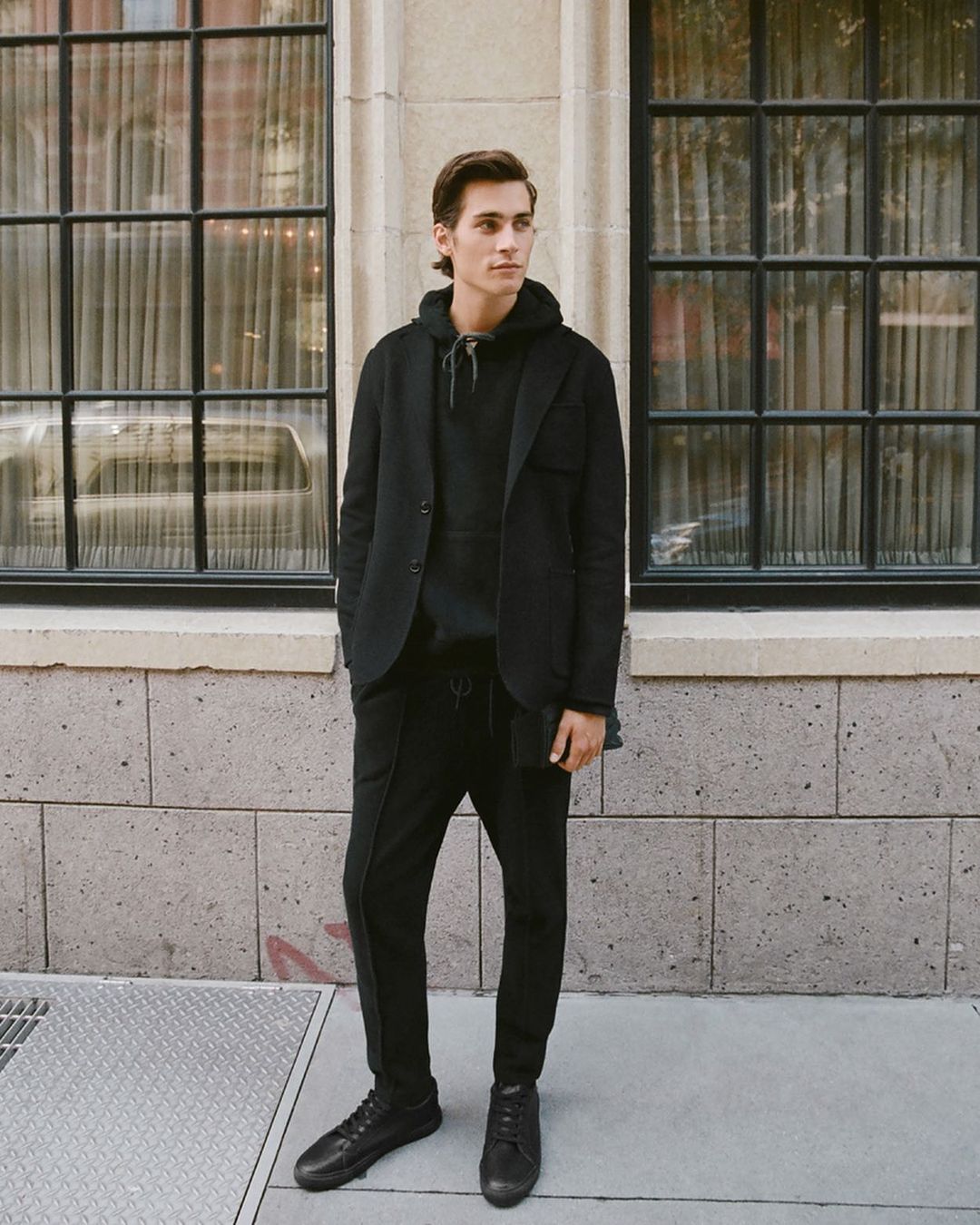 Full-length shot of a young adult man wearing a black blazer, hoodie, sweatpants and shoes standing outside a building with window.