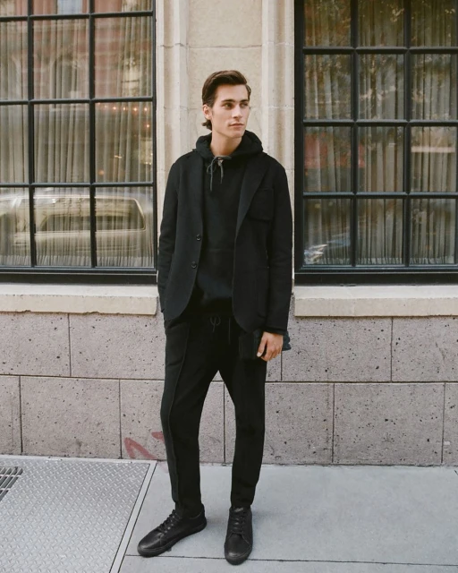 Full-length shot of a young adult man wearing a black blazer, hoodie, sweatpants and shoes standing outside a building with window.