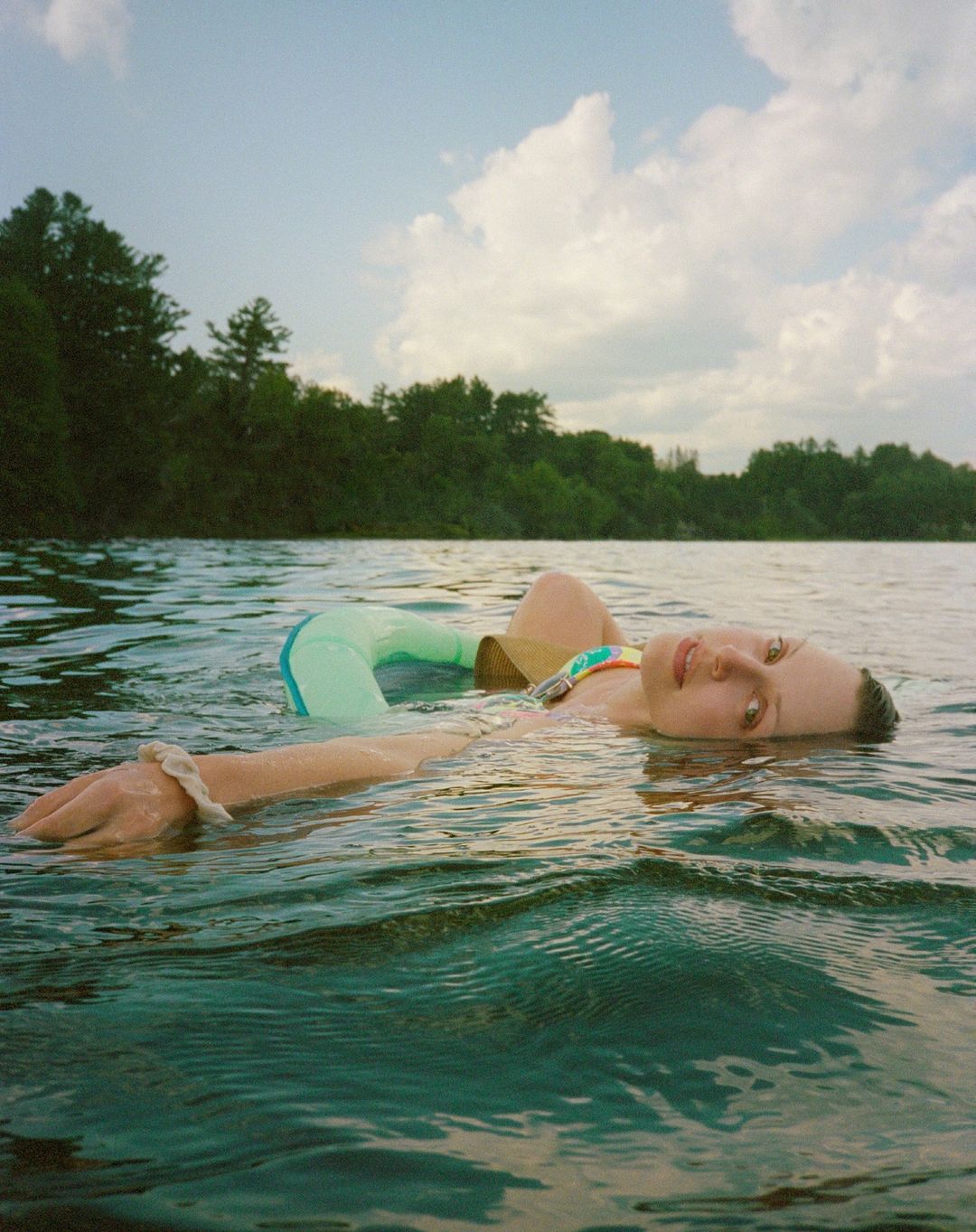 Woman floats on her back in rippling lake water, with trees and cloudy sky visible in the background. She wears a multi-colored swimsuit.