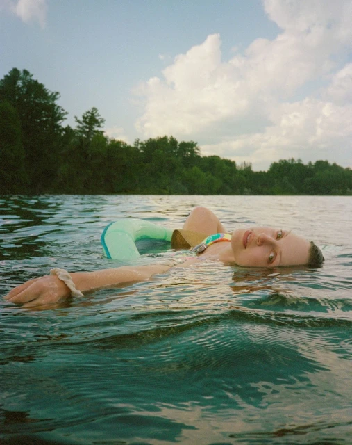 Woman floats on her back in rippling lake water, with trees and cloudy sky visible in the background. She wears a multi-colored swimsuit.