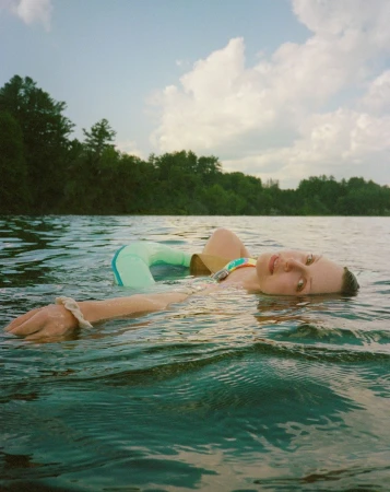 Woman floats on her back in rippling lake water, with trees and cloudy sky visible in the background. She wears a multi-colored swimsuit.