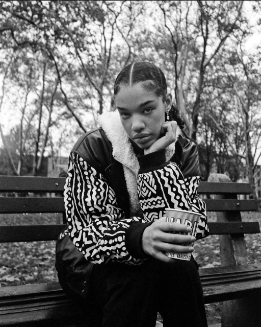Black and white portrait of a young woman with braided hair, seated on a park bench, holding a cup, and wearing a patterned jacket.