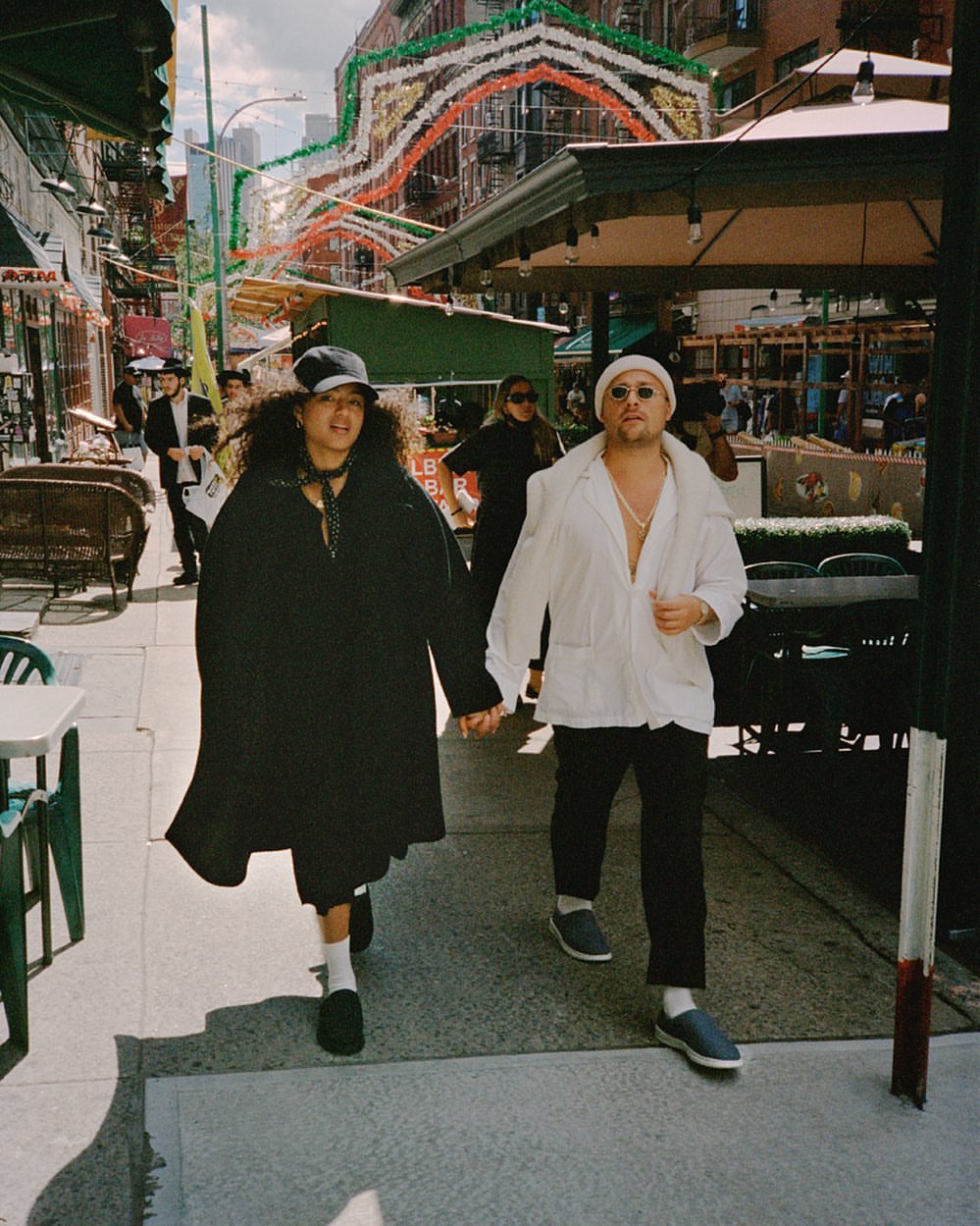 Couple walks hand-in-hand on sunny street with awnings, buildings, and hanging decorations in background. Street-style fashion.