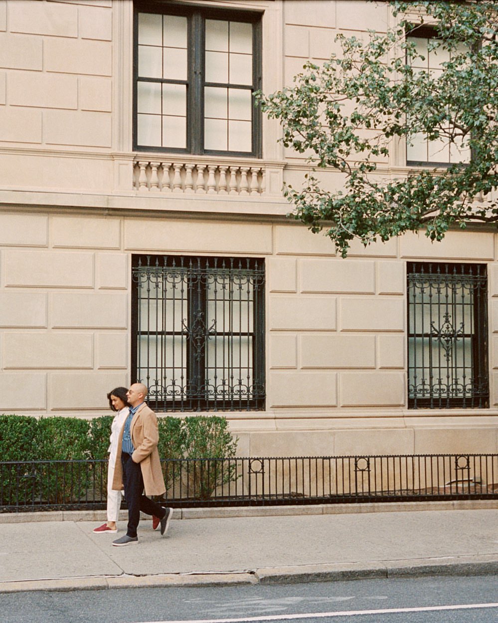 A man and woman walking together on a city sidewalk past a classic stone building facade with large windows.