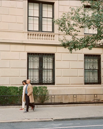 A man and woman walking together on a city sidewalk past a classic stone building facade with large windows.