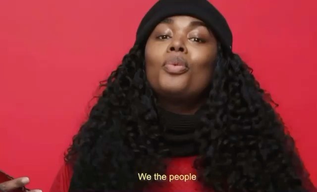 Portrait of a Black woman with curly hair and a beanie in front of a red background, looking at the camera, wearing a black beanie.