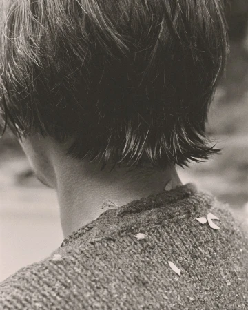 Close up shot of an adult with dark hair wearing a knit sweater with petals scattered across it, against a neutral backdrop.