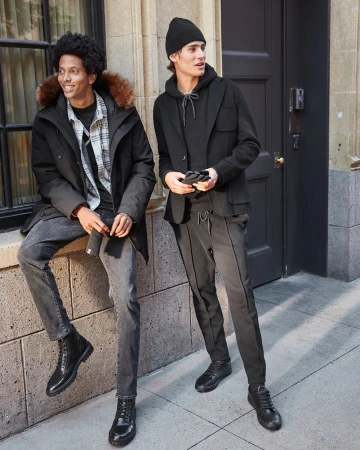Two adult men pose outdoors in urban setting, wearing jackets, sweaters, pants and boots in a fashion lifestyle product shot.
