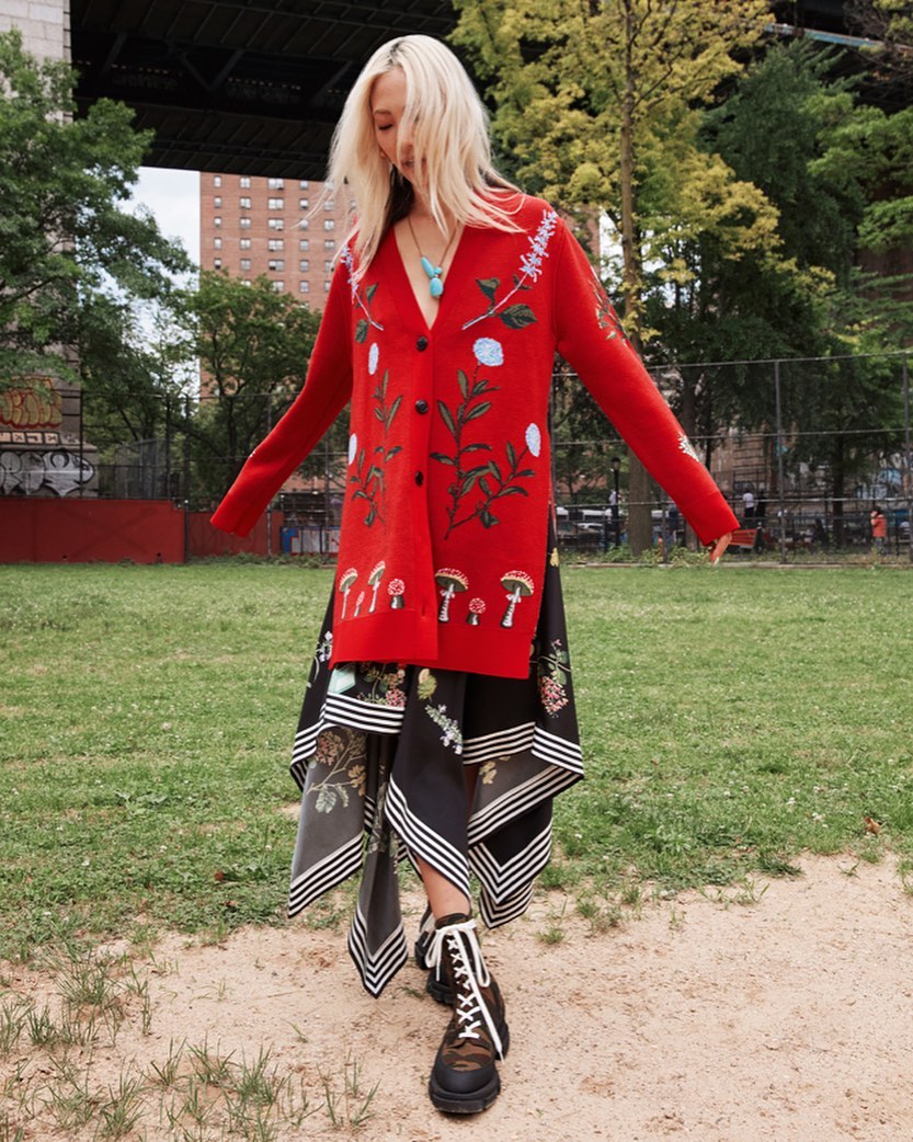 Woman in red floral cardigan and patterned skirt standing in a park with a bridge in the background; fashion editorial outdoor shot.