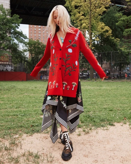 Woman in red floral cardigan and patterned skirt standing in a park with a bridge in the background; fashion editorial outdoor shot.