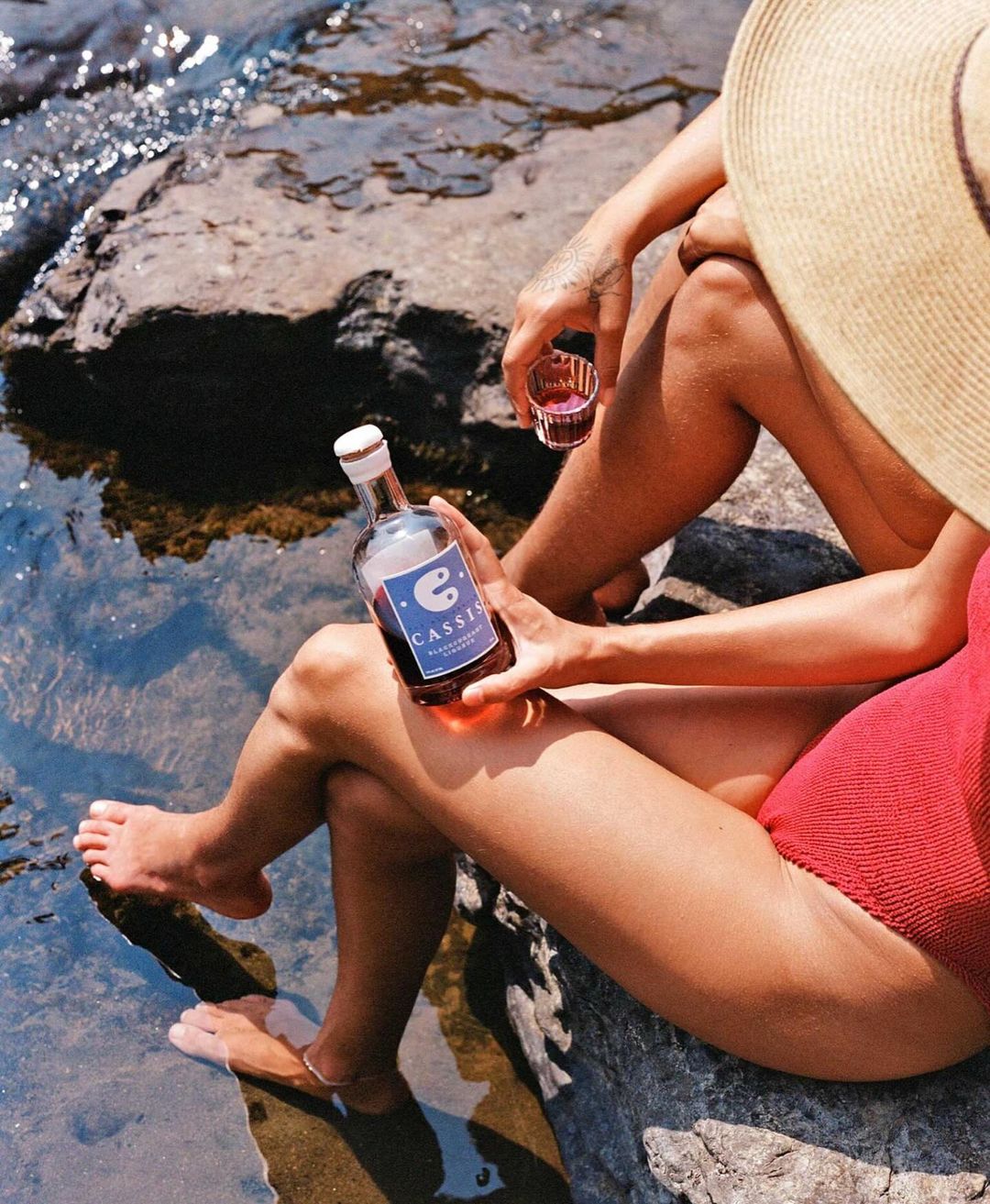 Two people on rocks by water, enjoying a bottle of Cassel Crianza wine in natural light, casual outdoor lifestyle, sunny day.